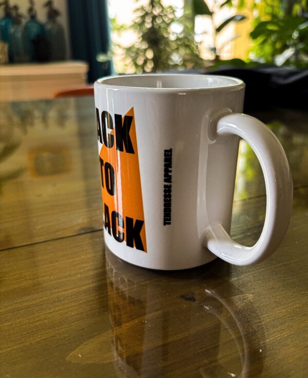 Photo d'un mug blanc posé sur une table en verre, sur le mug il est inscris Tendresse Apparel et sur le côté Back To Black en noir sur un triangle orange.