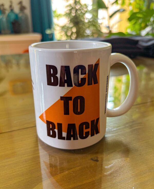 Photo d'un mug blanc posé sur une table en verre, sur le mug il est inscris Tendresse Apparel et sur le côté Back To Black en noir sur un triangle orange.