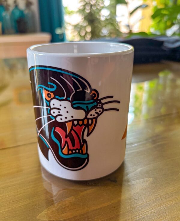 Une tasse blanche posée sur une table en verre présentant une panthère noir en motif tatouage traditionnel américain.