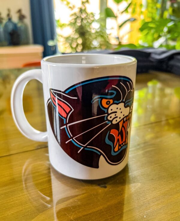 Une tasse blanche posée sur une table en verre présentant une panthère noir en motif tatouage traditionnel américain.