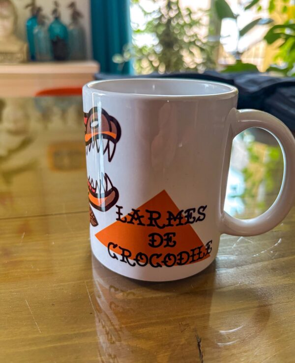 Une tasse blanche posée sur une table en verre présentant un crocodile qui pleure en motif tatouage traditionnel américain.