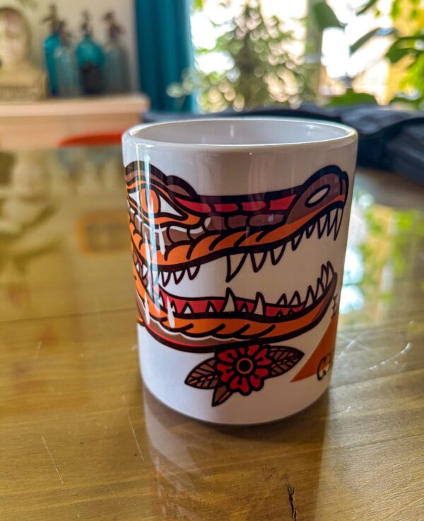 Une tasse blanche posée sur une table en verre présentant un crocodile qui pleure en motif tatouage traditionnel américain.