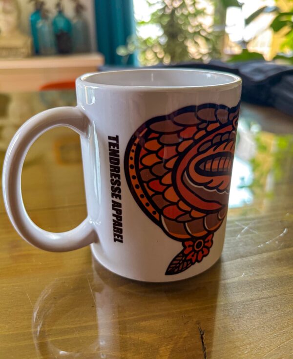 Une tasse blanche posée sur une table en verre présentant un crocodile qui pleure en motif tatouage traditionnel américain.
