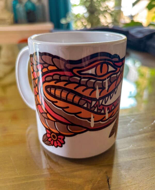 Une tasse blanche posée sur une table en verre présentant un crocodile qui pleure en motif tatouage traditionnel américain.