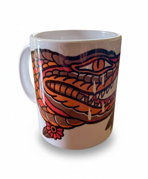 Une tasse blanche posée, présentant un crocodile qui pleure en motif tatouage traditionnel américain.