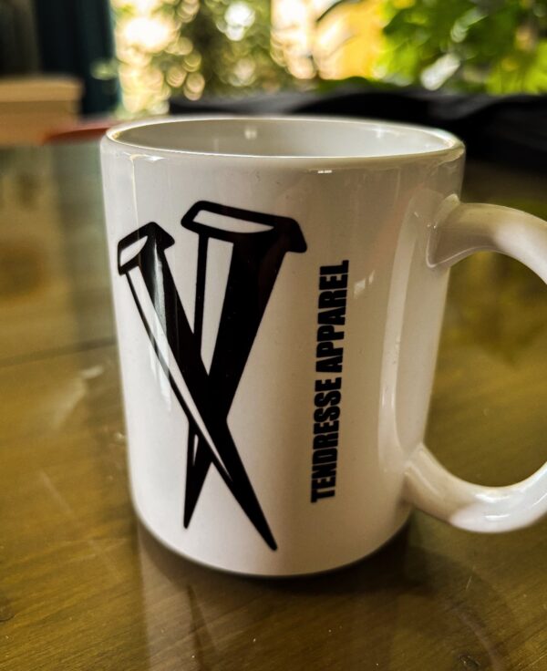 Une tasse blanche sur une table avec l'inscription Tendresse Apparel et 2 clous croisés en Noir.