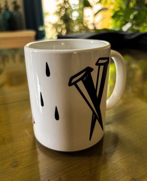 Une tasse blanche sur une table avec l'inscription Tendresse Apparel et 2 clous croisés en Noir.