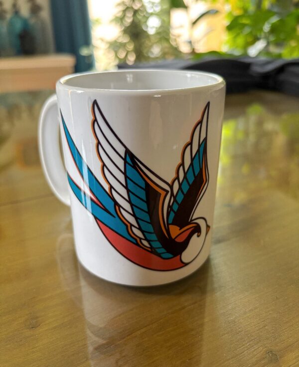 Une tasse blanche sur une table avec une hirondelle en motif tatouage traditionnel américain avec des larmes noires qui tombent.