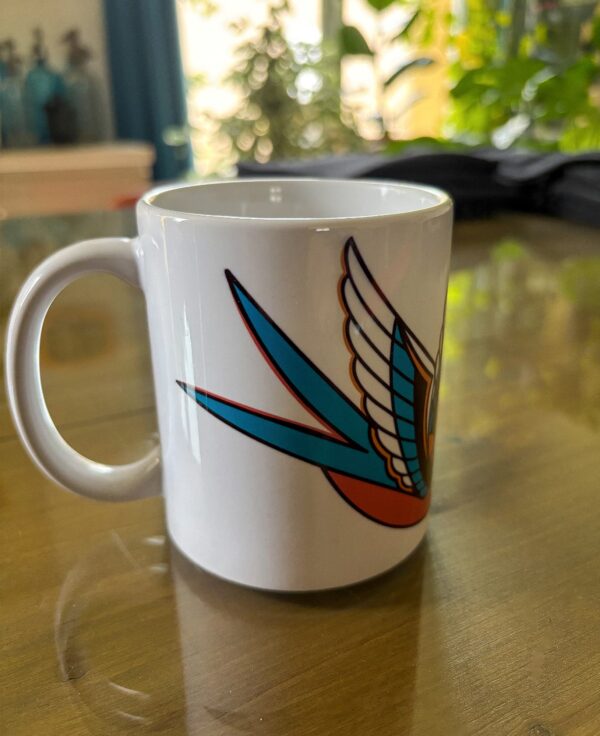 Une tasse blanche sur une table avec une hirondelle en motif tatouage traditionnel américain avec des larmes noires qui tombent.