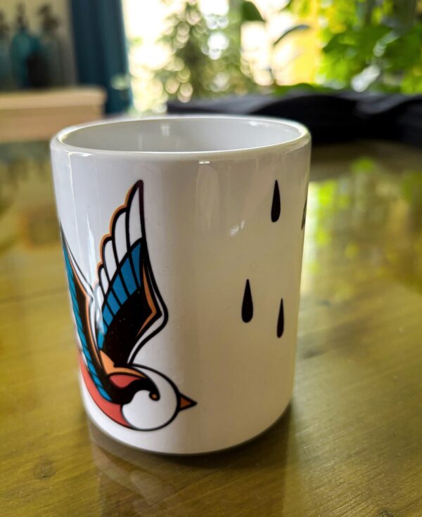 Une tasse blanche sur une table avec une hirondelle en motif tatouage traditionnel américain avec des larmes noires qui tombent.