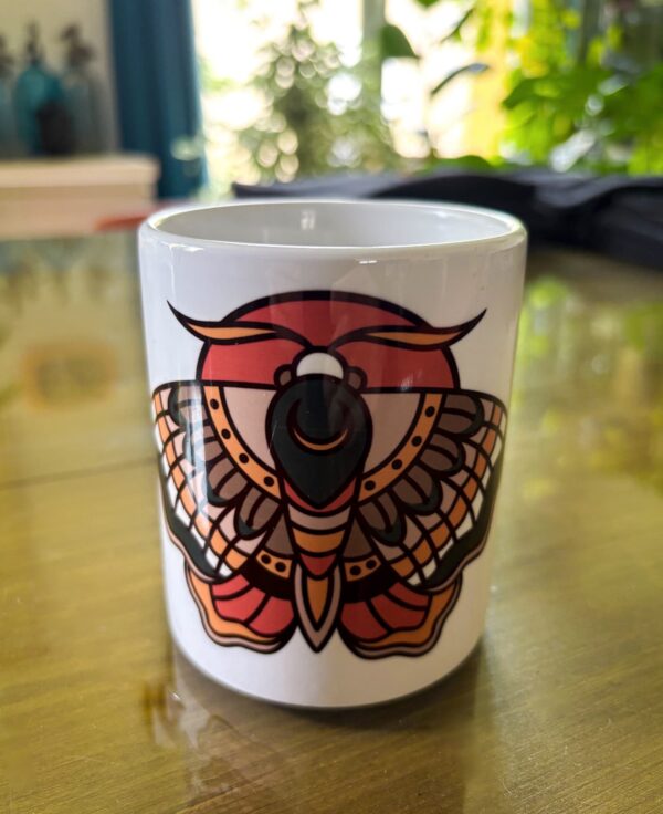 Une tasse blanche sur une table avec un papillon de nuit en motif tatouage traditionnel américain old school. Couleur Rouge, Brun, Vert, Jaune