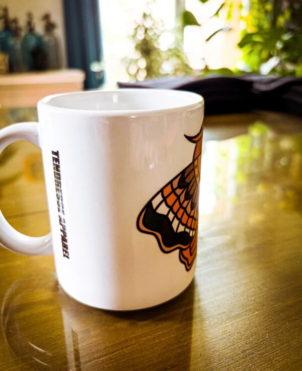 Une tasse blanche sur une table avec un papillon de nuit en motif tatouage traditionnel américain old school. Couleur Rouge, Brun, Vert, Jaune