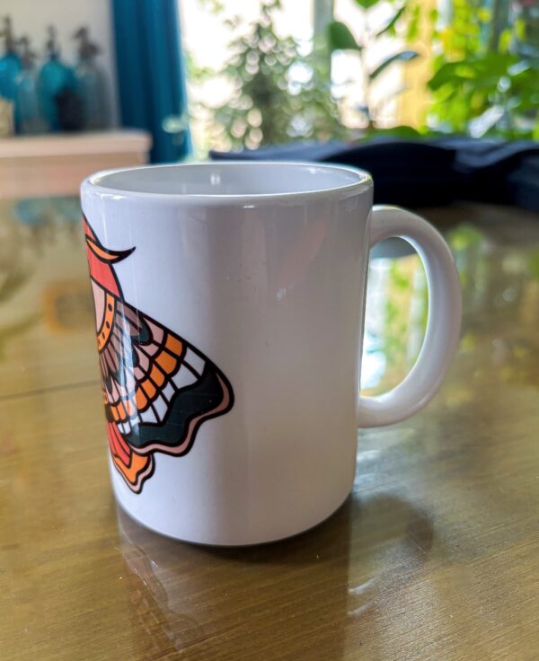 Une tasse blanche vu de côté, sur une table avec un papillon de nuit en motif tatouage traditionnel américain old school.